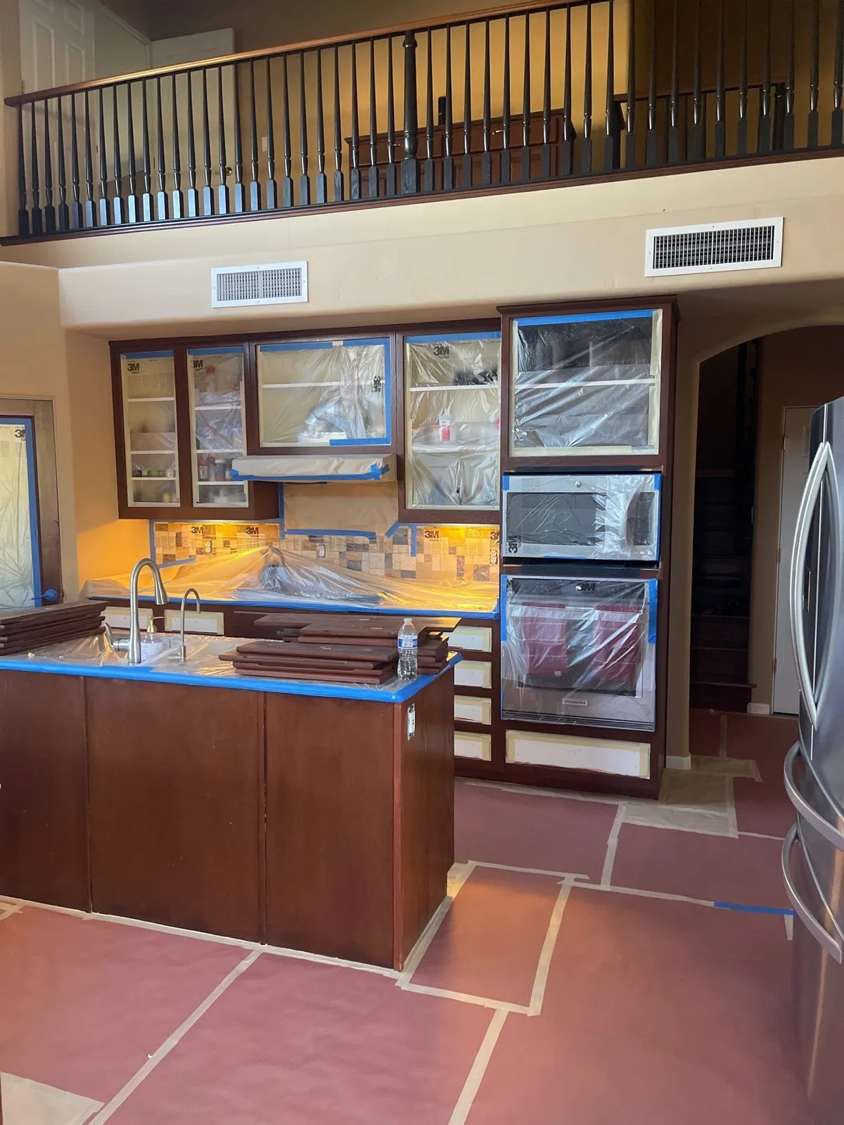 Dark-stained kitchen cabinets with glass-front uppers masked in plastic and blue tape, under a two-story ceiling with a balcony railing above