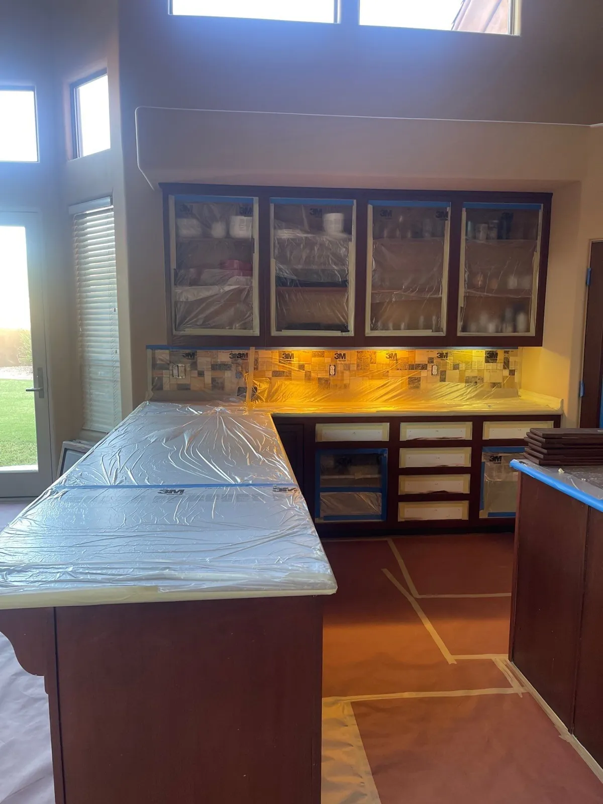 L-shaped dark-stained kitchen with countertops covered in plastic and floors covered in paper, ready for cabinet painting