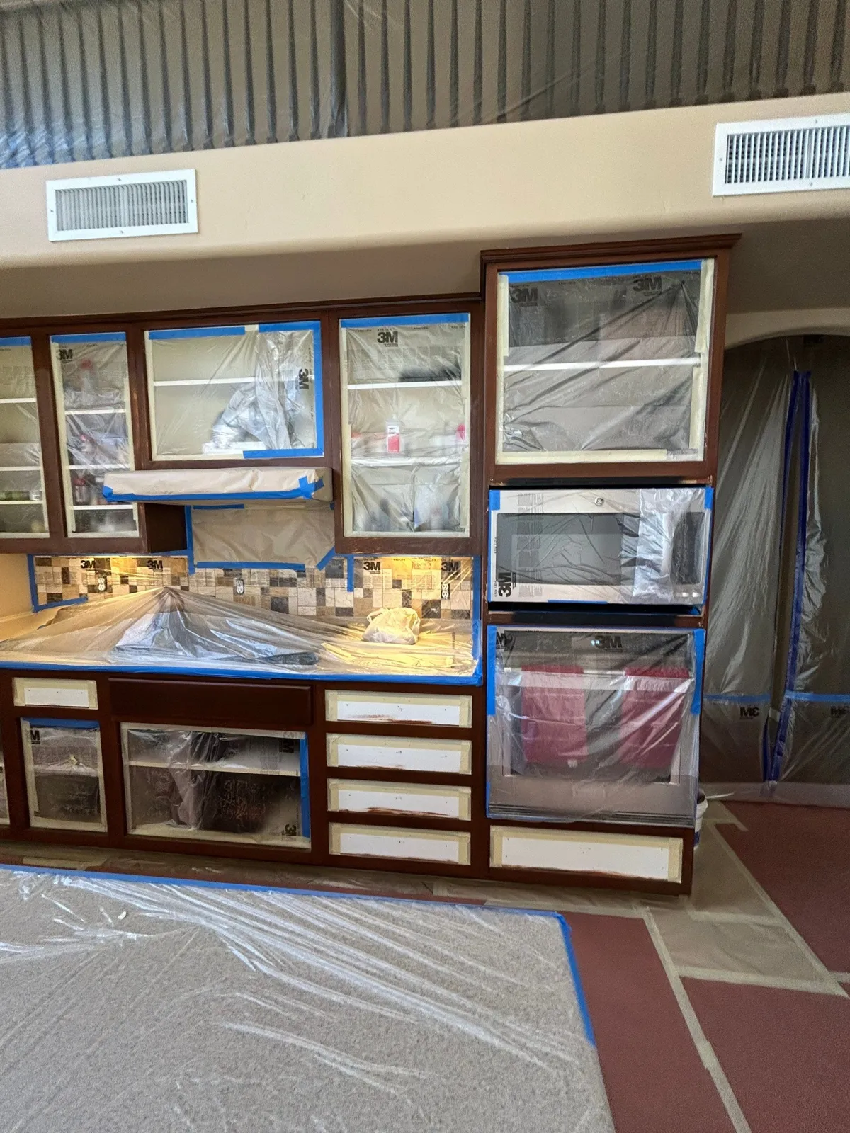 Wall of dark-stained cabinets and an oven tower, with appliances and glass doors sealed in plastic before painting