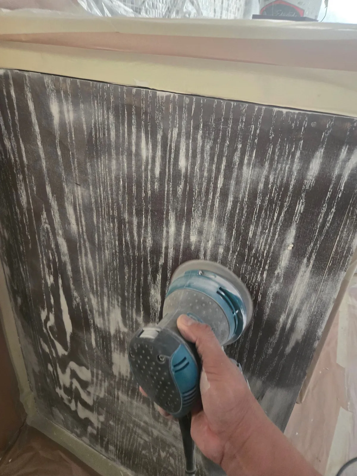 Orbital sander smoothing a dark cabinet panel, with white grain filler visible in the open wood grain