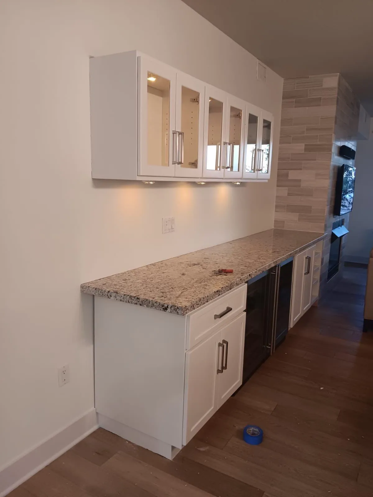 Bar wall with white glass-front upper cabinets and white shaker base cabinets along a granite counter